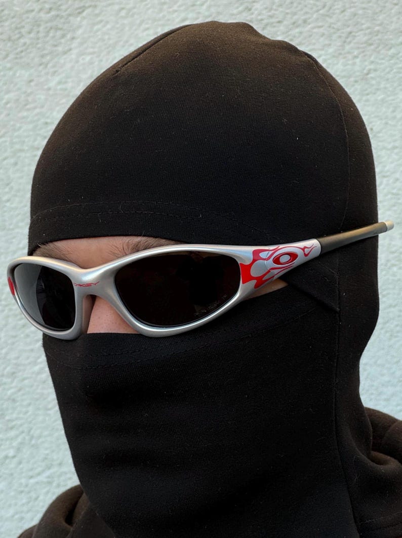 May include: Silver-framed sunglasses with red and white accents. The sunglasses have dark lenses and are worn with a black balaclava. The design on the frame is a flame-like pattern. The sunglasses are a sporty style.