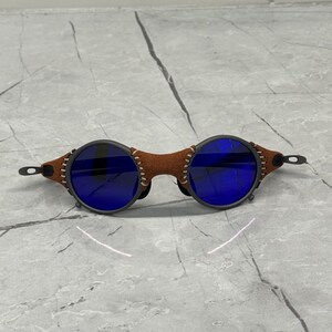 May include: Round sunglasses with dark gray frames and blue lenses. The bridge is brown leather with white stitching. The temples are thin and black. The sunglasses are set against a gray and white marble surface.