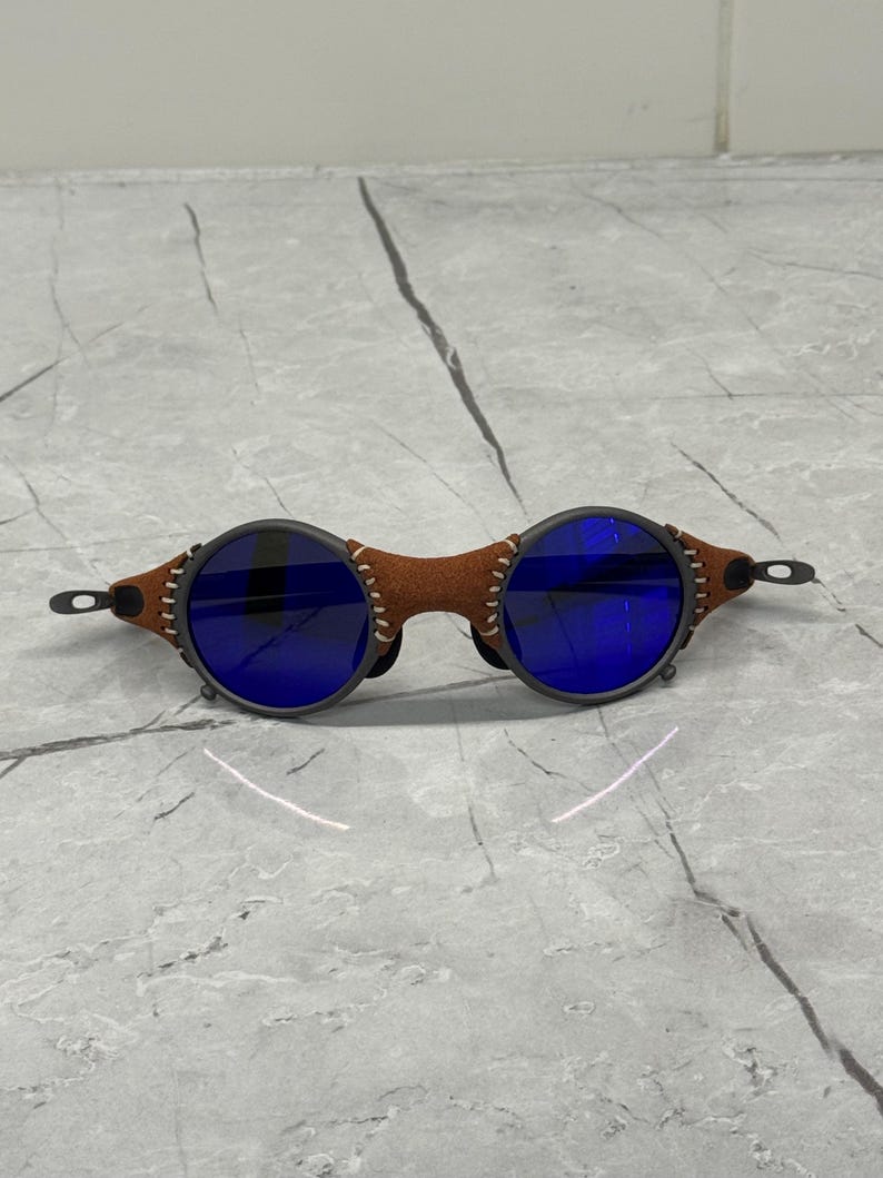 May include: Round sunglasses with dark gray frames and blue lenses. The bridge is brown leather with white stitching. The temples are thin and black. The sunglasses are set against a gray and white marble surface.