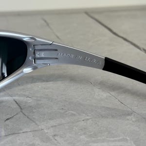 May include: Silver-framed sunglasses with dark lenses. The text "MADE IN U.S.A." is printed on the arm. The sunglasses have a sleek, modern design.