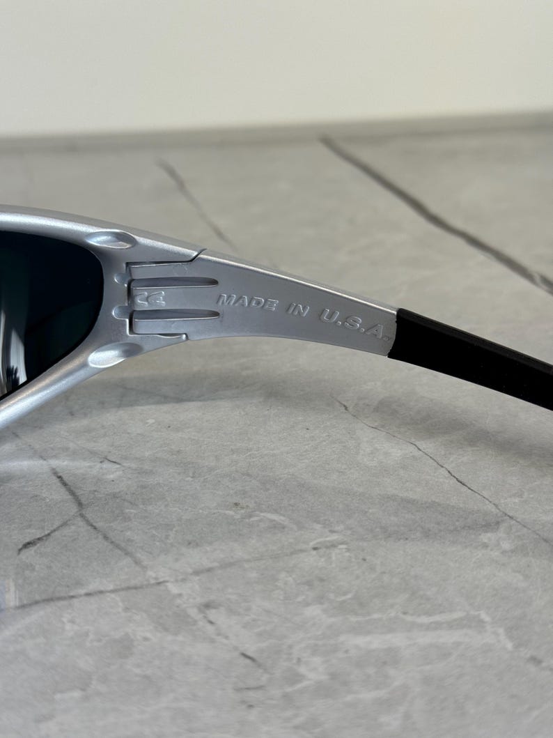May include: Silver-framed sunglasses with dark lenses. The text "MADE IN U.S.A." is printed on the arm. The sunglasses have a sleek, modern design.