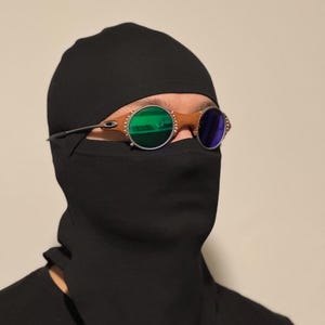 以下が含まれることがあります： 黒いバラクラバと黒いシャツを着た人。茶色のフレームと緑と青のレンズが付いた丸いサングラスをかけています。背景は無地でニュートラルな色です。