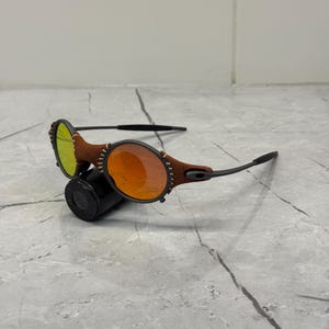 May include: Black framed sunglasses with orange and yellow lenses. The frames have brown leather accents with white stitching. The sunglasses are resting on a black cylindrical object against a gray marble surface.