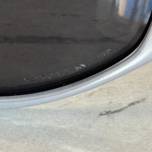 May include: Close-up of a silver-framed sunglass lens with the word "PRIZM" etched into the lens. The lens is dark, and the frame is a light silver color. The sunglasses are resting on a light gray surface.
