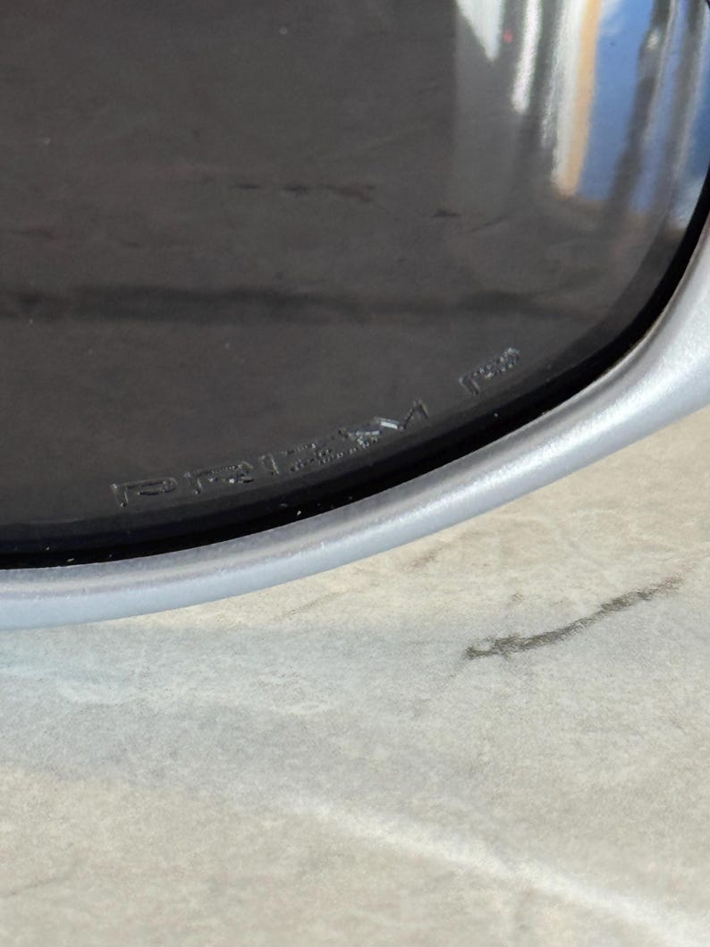 May include: Close-up of a silver-framed sunglass lens with the word "PRIZM" etched into the lens. The lens is dark, and the frame is a light silver color. The sunglasses are resting on a light gray surface.