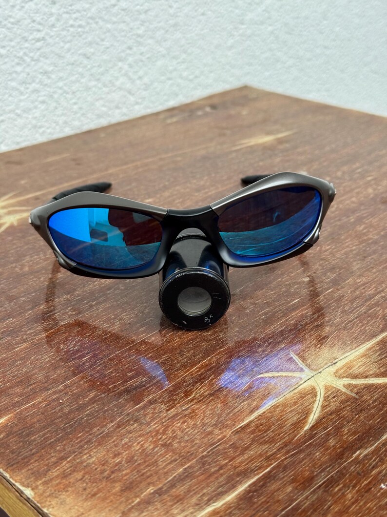 Op de afbeelding: Zwarte zonnebril met blauwe reflecterende glazen en een zwart montuur. De zonnebril ligt op een houten oppervlak. Een zwart apparaat met de tekst "8x" is eronder geplaatst.