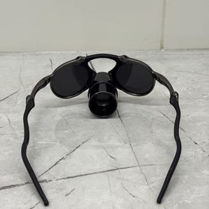 May include: Black sunglasses with a distinctive design. The sunglasses feature round lenses and a dark frame. A cylindrical attachment is present on the bridge. The arms of the sunglasses curve downwards.