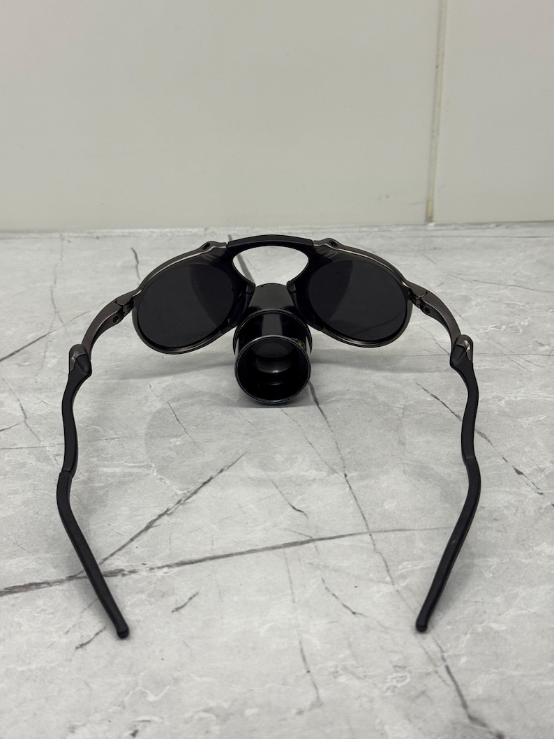 May include: Black sunglasses with a distinctive design. The sunglasses feature round lenses and a dark frame. A cylindrical attachment is present on the bridge. The arms of the sunglasses curve downwards.