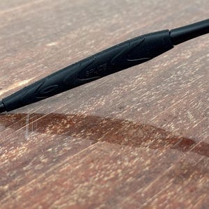 Op de afbeelding: Close-up van een zwarte brilpoot met het woord "SPLICE" erop gedrukt. De poot heeft een getextureerde grip. De achtergrond is een houten oppervlak.