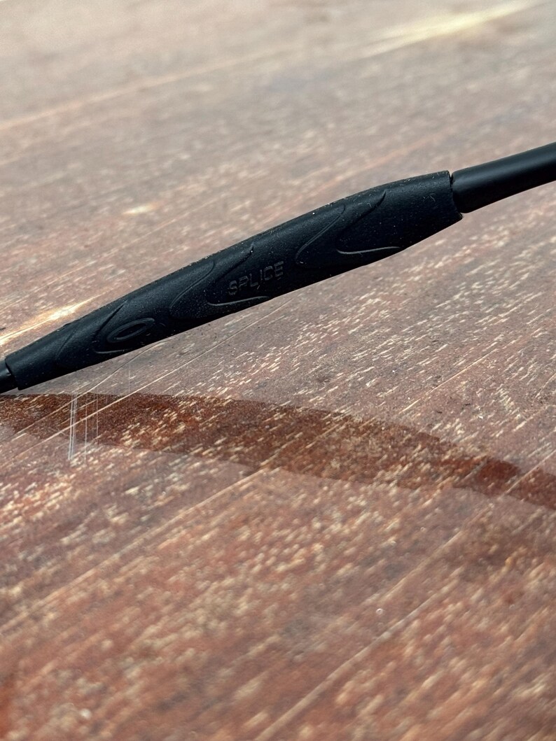 Op de afbeelding: Close-up van een zwarte brilpoot met het woord "SPLICE" erop gedrukt. De poot heeft een getextureerde grip. De achtergrond is een houten oppervlak.