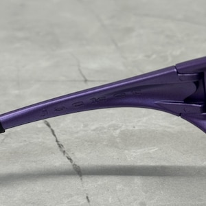 May include: Close-up of a pair of purple sunglasses. The word "jacket" is printed on the arm of the sunglasses. The sunglasses are on a gray surface.