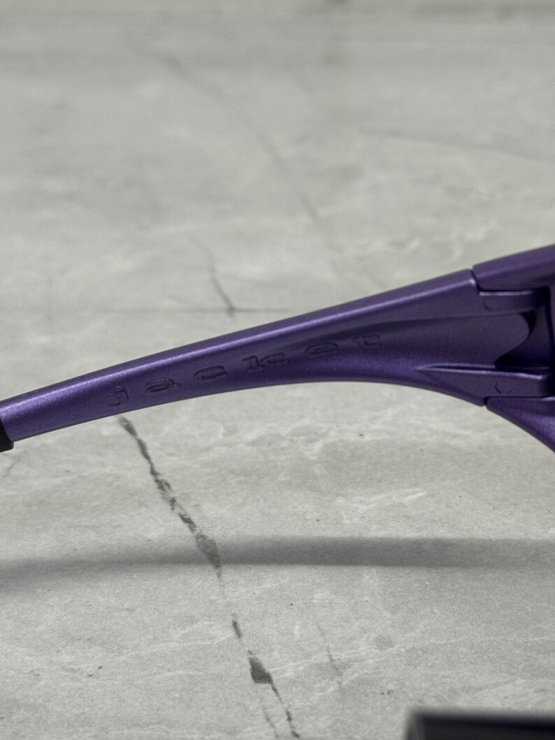 May include: Close-up of a pair of purple sunglasses. The word "jacket" is printed on the arm of the sunglasses. The sunglasses are on a gray surface.