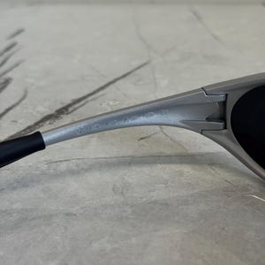 Puede incluir: Primer plano de unas gafas de sol grises con lentes negras. Las gafas de sol tienen un dise&ntilde;o elegante y moderno con el nombre de la marca grabado en la patilla. La montura es de color gris mate.