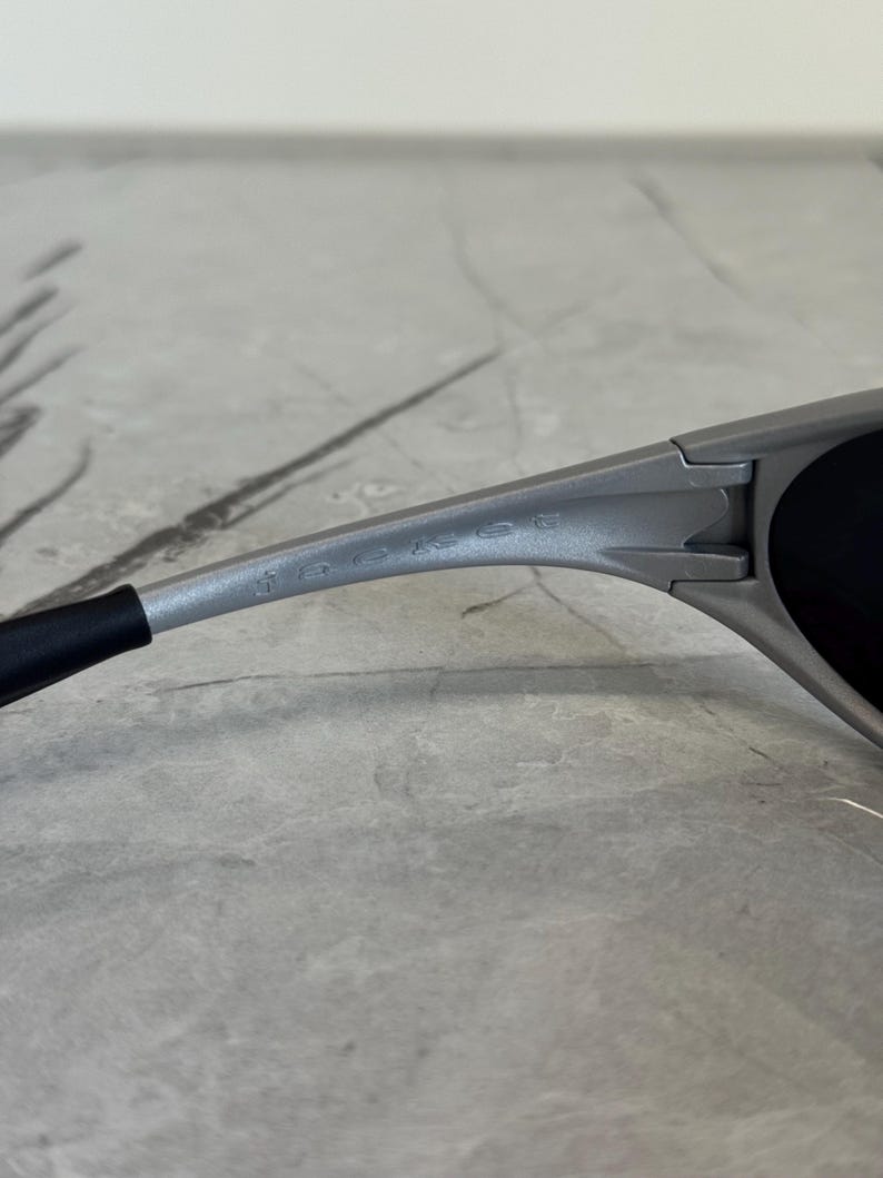 Puede incluir: Primer plano de unas gafas de sol grises con lentes negras. Las gafas de sol tienen un dise&ntilde;o elegante y moderno con el nombre de la marca grabado en la patilla. La montura es de color gris mate.