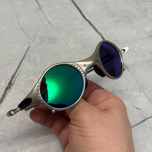 Może przedstawiać: Srebrne okulary przeciwsłoneczne z okrągłymi zielonymi soczewkami i niebieską soczewką. Okulary przeciwsłoneczne mają unikalny design z mostkiem i zausznikami. Soczewki odbijają światło, a oprawka ma metaliczne wykończenie.