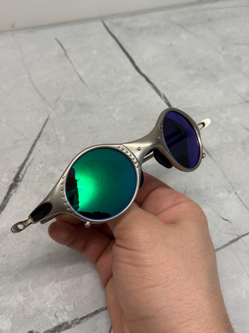 Może przedstawiać: Srebrne okulary przeciwsłoneczne z okrągłymi zielonymi soczewkami i niebieską soczewką. Okulary przeciwsłoneczne mają unikalny design z mostkiem i zausznikami. Soczewki odbijają światło, a oprawka ma metaliczne wykończenie.