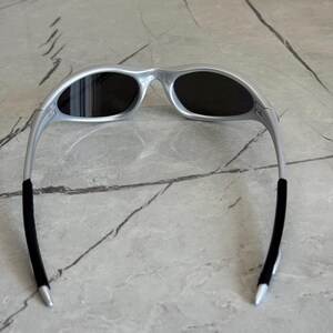 May include: Silver sunglasses with dark lenses. The sunglasses have a sleek, modern design with a curved frame and black rubber temple tips. The oval lenses offer full coverage. The frame is made of a lightweight material.