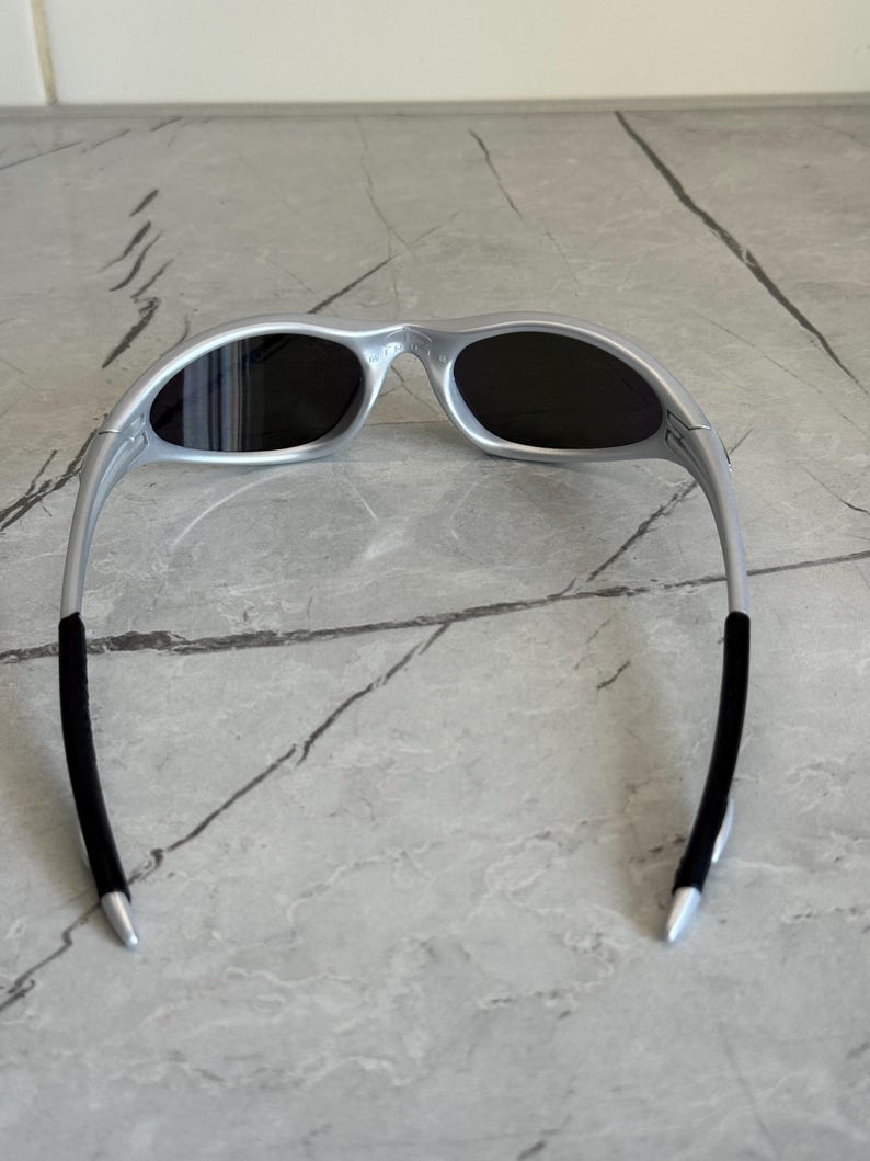 May include: Silver sunglasses with dark lenses. The sunglasses have a sleek, modern design with a curved frame and black rubber temple tips. The oval lenses offer full coverage. The frame is made of a lightweight material.