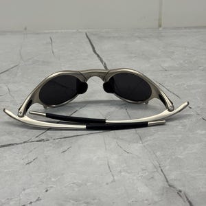 May include: Silver-framed sunglasses with dark lenses. The sunglasses have a sleek, modern design with a curved frame and black temple arms. The lenses are oval-shaped and appear to be polarized.