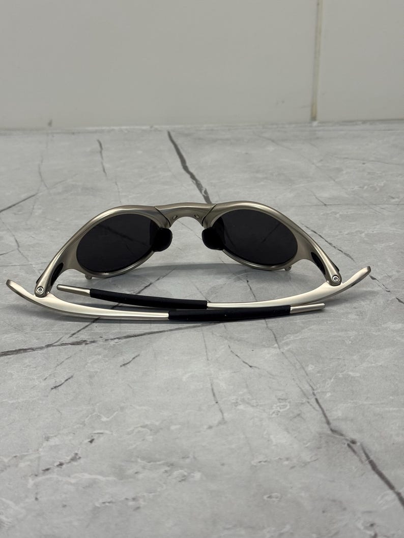 May include: Silver-framed sunglasses with dark lenses. The sunglasses have a sleek, modern design with a curved frame and black temple arms. The lenses are oval-shaped and appear to be polarized.