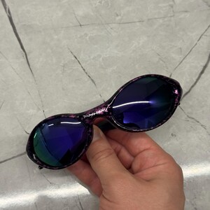 May include: Oval sunglasses with a speckled purple frame and dark blue lenses. The lenses reflect a green hue. The sunglasses are held by a hand.