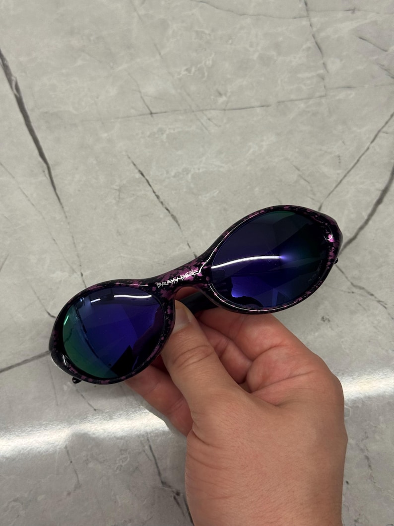 May include: Oval sunglasses with a speckled purple frame and dark blue lenses. The lenses reflect a green hue. The sunglasses are held by a hand.