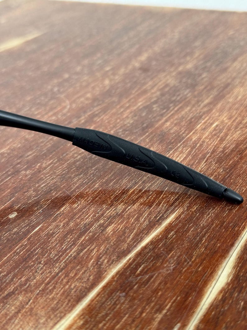 Op de afbeelding: Close-up van een zwarte brilpoot met de tekst "MADE IN USA" en het nummer "C3" erop gedrukt. De poot heeft een getextureerde grip en ligt op een houten oppervlak.