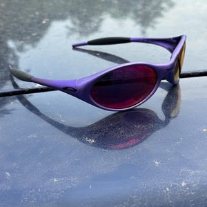 May include: Purple sunglasses with a sleek, modern design. The frames are a vibrant purple, complemented by dark lenses. The arms have black accents for added contrast. The sunglasses are designed for eye protection and fashion.