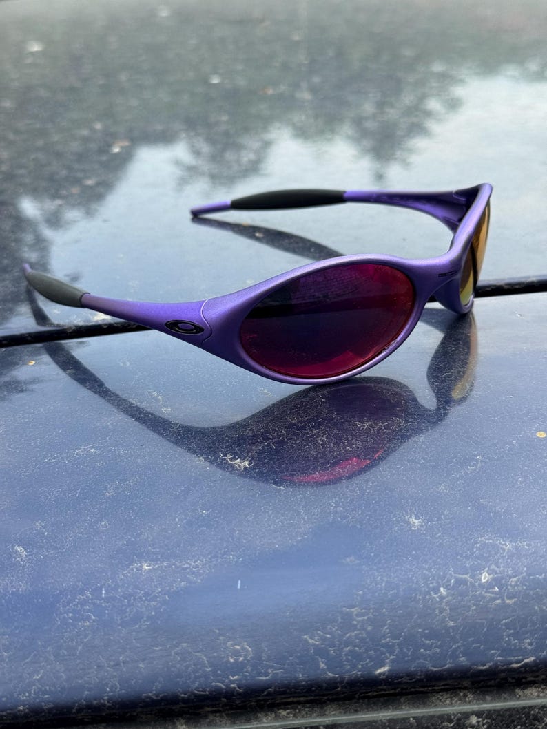 May include: Purple sunglasses with a sleek, modern design. The frames are a vibrant purple, complemented by dark lenses. The arms have black accents for added contrast. The sunglasses are designed for eye protection and fashion.