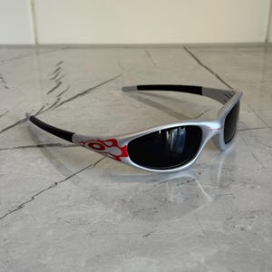 May include: Silver-framed sunglasses with black lenses and temples. The frame features a red and orange flame design on the side. The sunglasses are designed for eye protection and fashion.