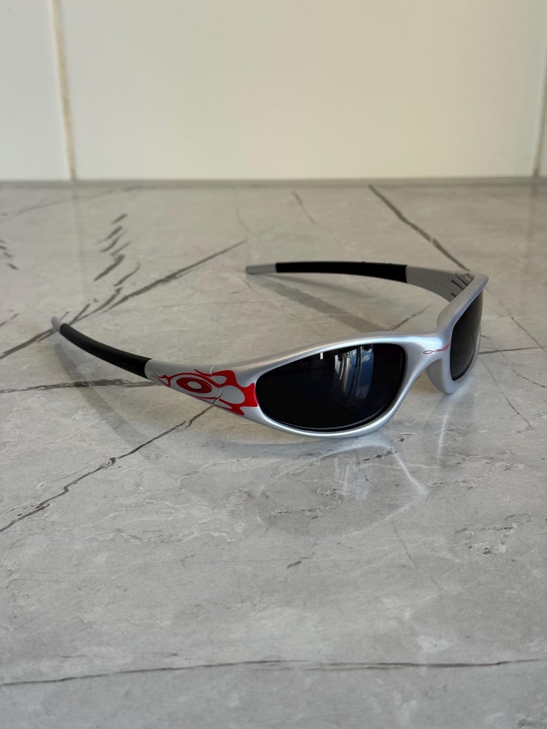 May include: Silver-framed sunglasses with black lenses and temples. The frame features a red and orange flame design on the side. The sunglasses are designed for eye protection and fashion.