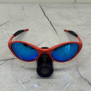 Op de afbeelding: Rood omrande zonnebril met blauwe reflecterende glazen. De zonnebril heeft een gebogen ontwerp en zwarte accenten op de veren. De glazen reflecteren de omgeving. De zonnebril rust op een zwart cilindrisch object.