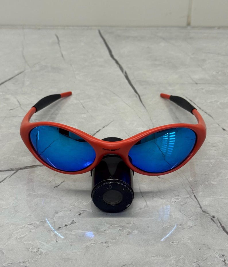Op de afbeelding: Rood omrande zonnebril met blauwe reflecterende glazen. De zonnebril heeft een gebogen ontwerp en zwarte accenten op de veren. De glazen reflecteren de omgeving. De zonnebril rust op een zwart cilindrisch object.
