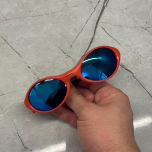 Op de afbeelding: Rode zonnebril met blauwe spiegelglazen. De zonnebril heeft een gebogen montuurontwerp. De glazen reflecteren de omgeving. De zonnebril wordt door een hand vastgehouden.