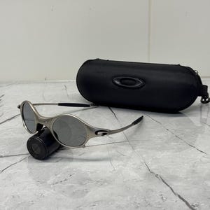 May include: Silver-framed sunglasses with gray lenses and a black case. The sunglasses have a sleek, modern design with a curved frame and a small logo on the temple. The black case is oval-shaped with a zipper closure.