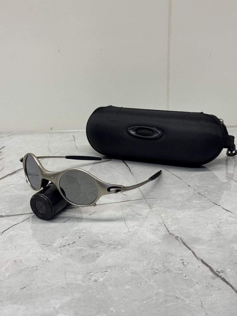 May include: Silver-framed sunglasses with gray lenses and a black case. The sunglasses have a sleek, modern design with a curved frame and a small logo on the temple. The black case is oval-shaped with a zipper closure.