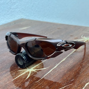 Puede incluir: Gafas de sol marrones con montura met&aacute;lica y lentes oscuras. Las gafas tienen un dise&ntilde;o elegante y moderno con forma curvada y un peque&ntilde;o logotipo en las patillas. Las lentes son de color marr&oacute;n y la montura es marr&oacute;n.