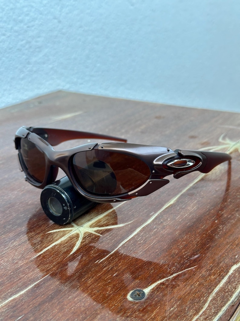 Puede incluir: Gafas de sol marrones con montura met&aacute;lica y lentes oscuras. Las gafas tienen un dise&ntilde;o elegante y moderno con forma curvada y un peque&ntilde;o logotipo en las patillas. Las lentes son de color marr&oacute;n y la montura es marr&oacute;n.
