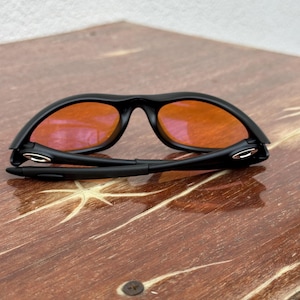 May include: Black sunglasses with orange lenses are folded on a wooden surface. The sunglasses have a sleek, modern design with a matte black frame. The lenses are a vibrant orange color, contrasting with the black frame. The wooden surface has a natural wood grain pattern.