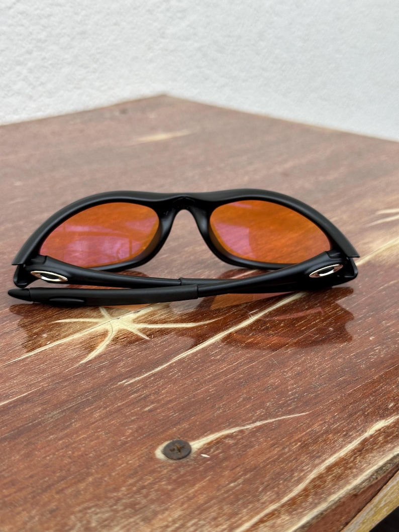 May include: Black sunglasses with orange lenses are folded on a wooden surface. The sunglasses have a sleek, modern design with a matte black frame. The lenses are a vibrant orange color, contrasting with the black frame. The wooden surface has a natural wood grain pattern.