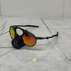 Pu&ograve; includere: Occhiali da sole neri con lenti arancioni e gialle. Gli occhiali hanno un design unico con una lente circolare e una montatura curva. Le lenti presentano un effetto sfumato, che passa dall'arancione al giallo. Gli occhiali sono appoggiati su una superficie grigia.