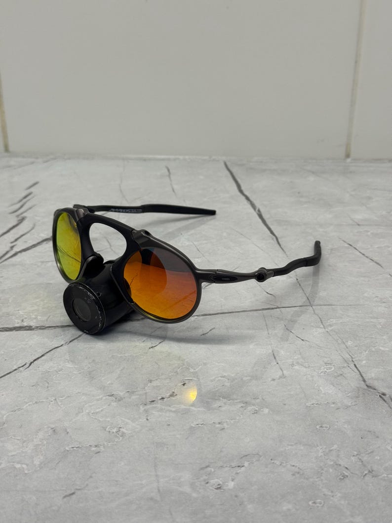 Pu&ograve; includere: Occhiali da sole neri con lenti arancioni e gialle. Gli occhiali hanno un design unico con una lente circolare e una montatura curva. Le lenti presentano un effetto sfumato, che passa dall'arancione al giallo. Gli occhiali sono appoggiati su una superficie grigia.