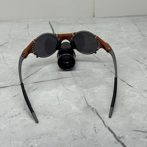 May include: Round sunglasses with dark lenses and a brown leather bridge. The frames are a dark gray color, with brown leather accents on the sides. The sunglasses have a unique, vintage style.