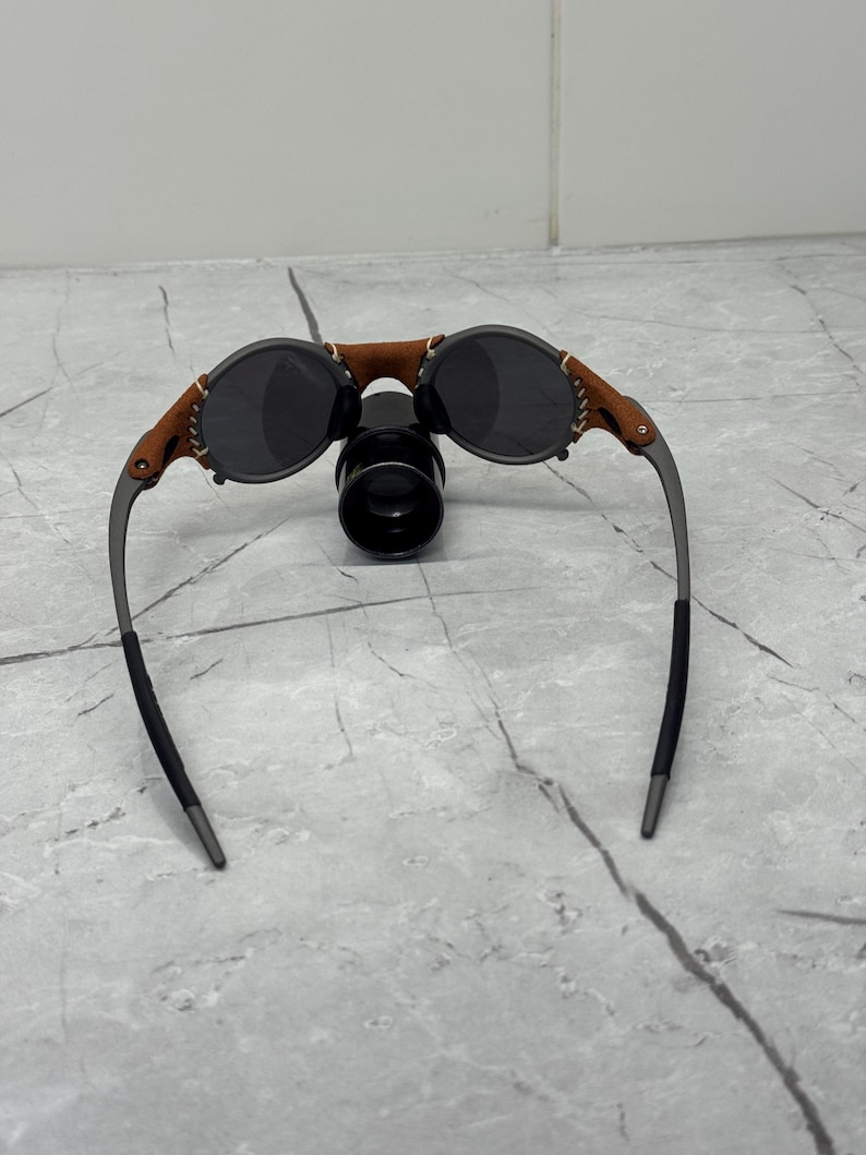 May include: Round sunglasses with dark lenses and a brown leather bridge. The frames are a dark gray color, with brown leather accents on the sides. The sunglasses have a unique, vintage style.