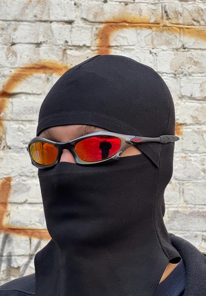Op de afbeelding: Zilverkleurige en zwarte zonnebril met oranje getinte glazen, gedragen met een zwarte bivakmuts. De zonnebril heeft een strak, sportief design. De achtergrond is een verweerde bakstenen muur met oranje vlekken.
