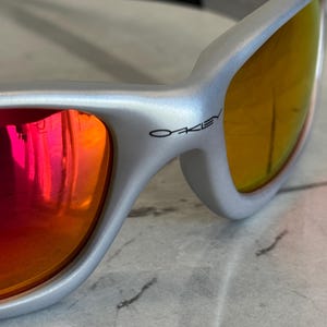 May include: Silver-framed sunglasses with orange and red mirrored lenses. The brand name "Oakley" is printed in black on the temple. The sunglasses have a sleek, modern design.