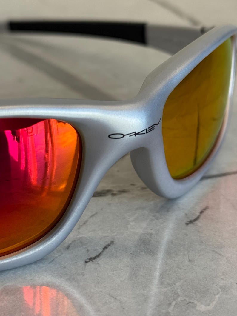 May include: Silver-framed sunglasses with orange and red mirrored lenses. The brand name "Oakley" is printed in black on the temple. The sunglasses have a sleek, modern design.