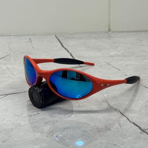 Op de afbeelding: Rode zonnebril met blauwe spiegelglazen. De zonnebril heeft zwarte oorpootjes en ligt op een zwart cilindrisch object. De glazen reflecteren de omgeving.