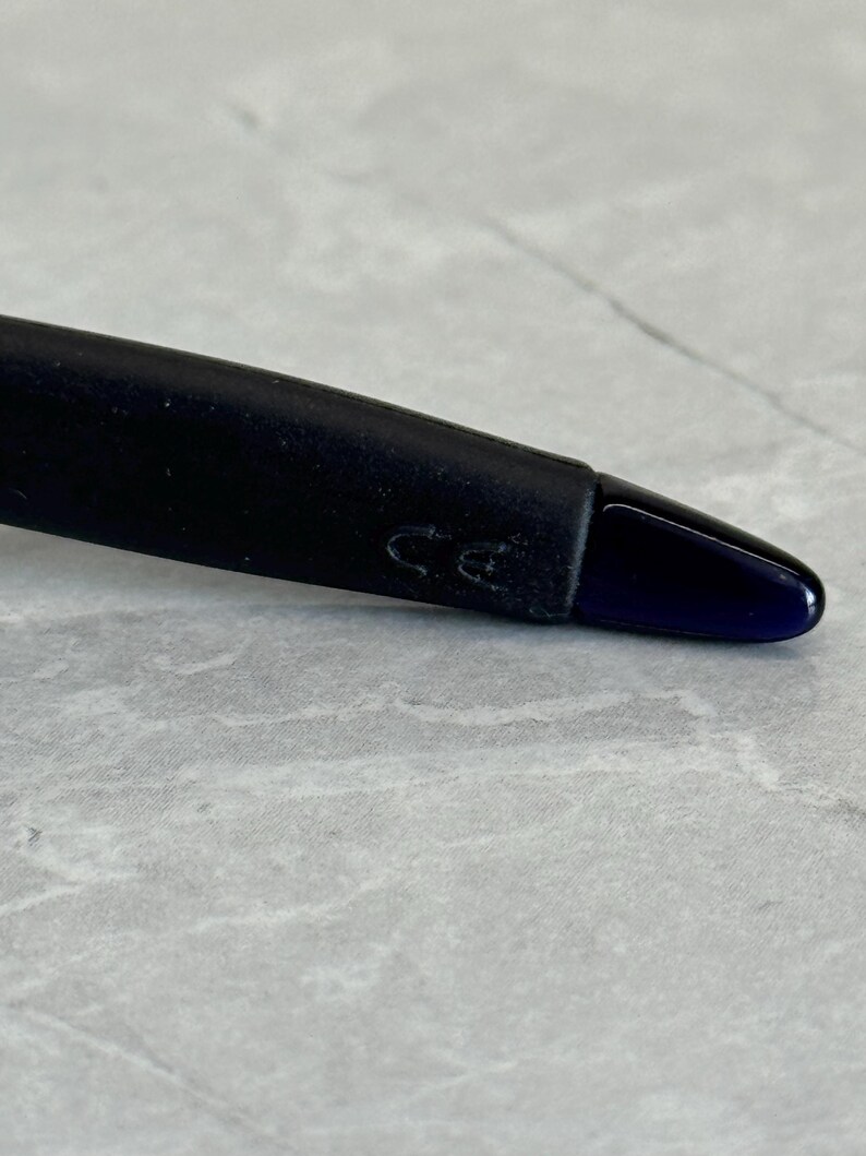 May include: Close-up of a black pen with a dark blue tip. The pen's body is black and has a matte finish. The pen's tip is a glossy dark blue color. The pen is resting on a gray and white marble surface.