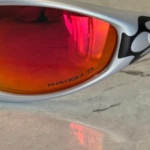 May include: Silver-framed sunglasses with red-orange lenses. The lenses have the text "PRIZM P" printed on them. The frame has a black and white design on the temple.